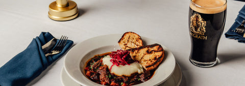Dish with bread and sauce next to a glass of dark House of McGrath beer on a white tablecloth.

