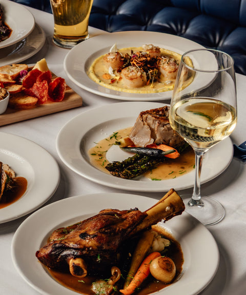Table with various dishes of food and drinks, including plates of meat and vegetables, a glass of white wine, and a beer.