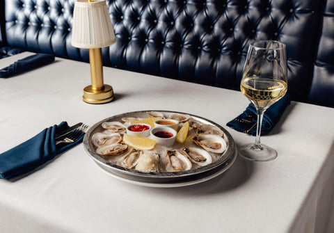 Platter of oysters with a glass of white wine on a table with a dark leather couch in the background.