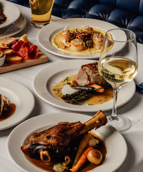 Table with various dishes of food and drinks, including plates of meat and vegetables, a glass of white wine, and house-brewed beer.