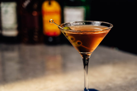 Martini glass with a cocktail and olives on a bar counter, with blurred bottles in the background.