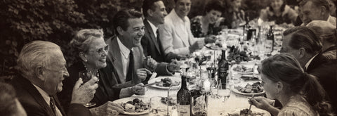 A black and white photograph depicting a long dinner table filled with people in an outdoor setting.